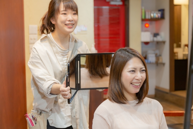 ヘアメイクの仕上がりを合わせ鏡で、笑顔で見せる美容師。客も満足して笑っている。
