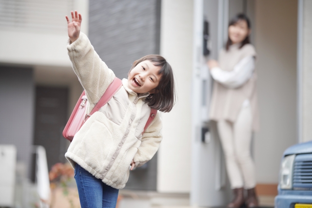 元気に手を振るランドセルを背負った女の子。後ろには玄関で笑顔の母親が見守っている。